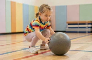 Petite fille en tenue de sport qui donne un coup de pied dans un ballon en mousse dans une salle de gym colorée